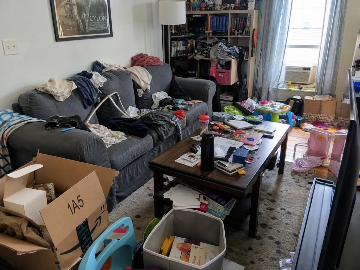 Sala bagunçada com roupas sobre o sofá, caixas de papelão e brinquedos no chão